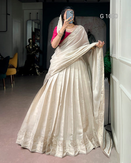 A woman takes a mirror selfie wearing a traditional Indian lehenga choli. The lehenga is a light beige/cream color with subtle gold embroidery along the border, paired with a bright pink sleeveless blouse.