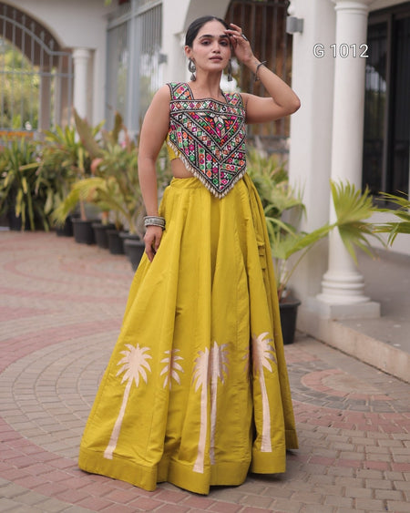 A woman models a traditional Indian outfit featuring a bright yellow flared skirt with white palm tree prints and a heavily embroidered sleeveless top, standing on a brick walkway in front of a building with white pillars.