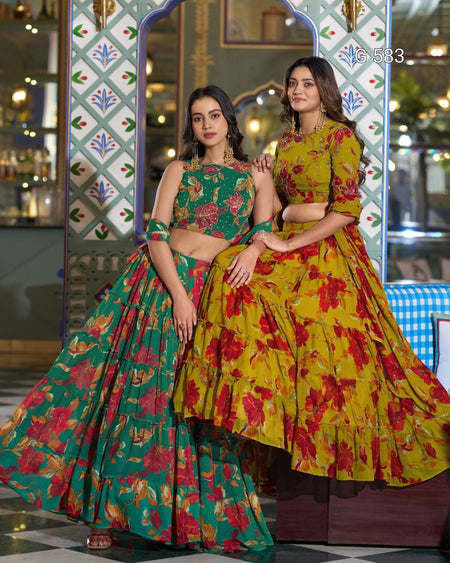 Two women modeling traditional Indian lehenga choli suits in a decorated indoor setting. The woman on the left wears a green floral lehenga with a matching green choli, and the woman on the right wears a yellow floral lehenga with a matching yellow choli.