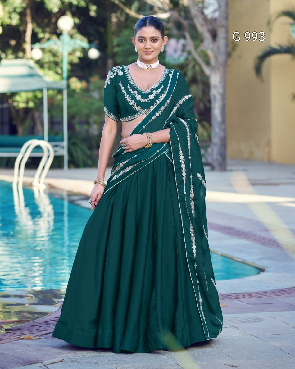 A woman stands outdoors beside a swimming pool wearing a dark green traditional outfit with silver embellished embroidery and a matching draped scarf. The background includes trees, a patio, and pool water.