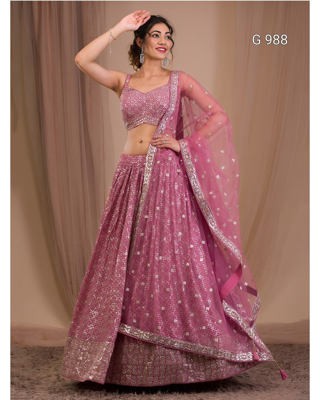 A woman modeling a pink embellished lehenga choli set with a matching dupatta, standing against a beige curtain backdrop. The outfit features silver mirror work and sequin embellishments, and the text 'G 988' is visible in the top right corner.
