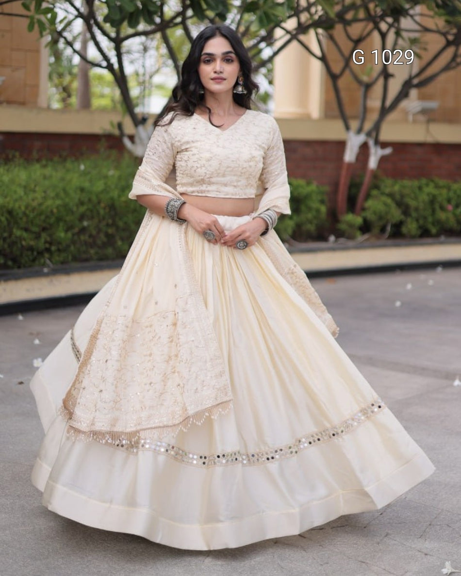 A woman modeling a cream-colored traditional Indian lehenga choli set outdoors. The set features a heavily embroidered crop blouse with three-quarter sleeves, a matching flared skirt with lace panels, and a dupatta draped over one shoulder.