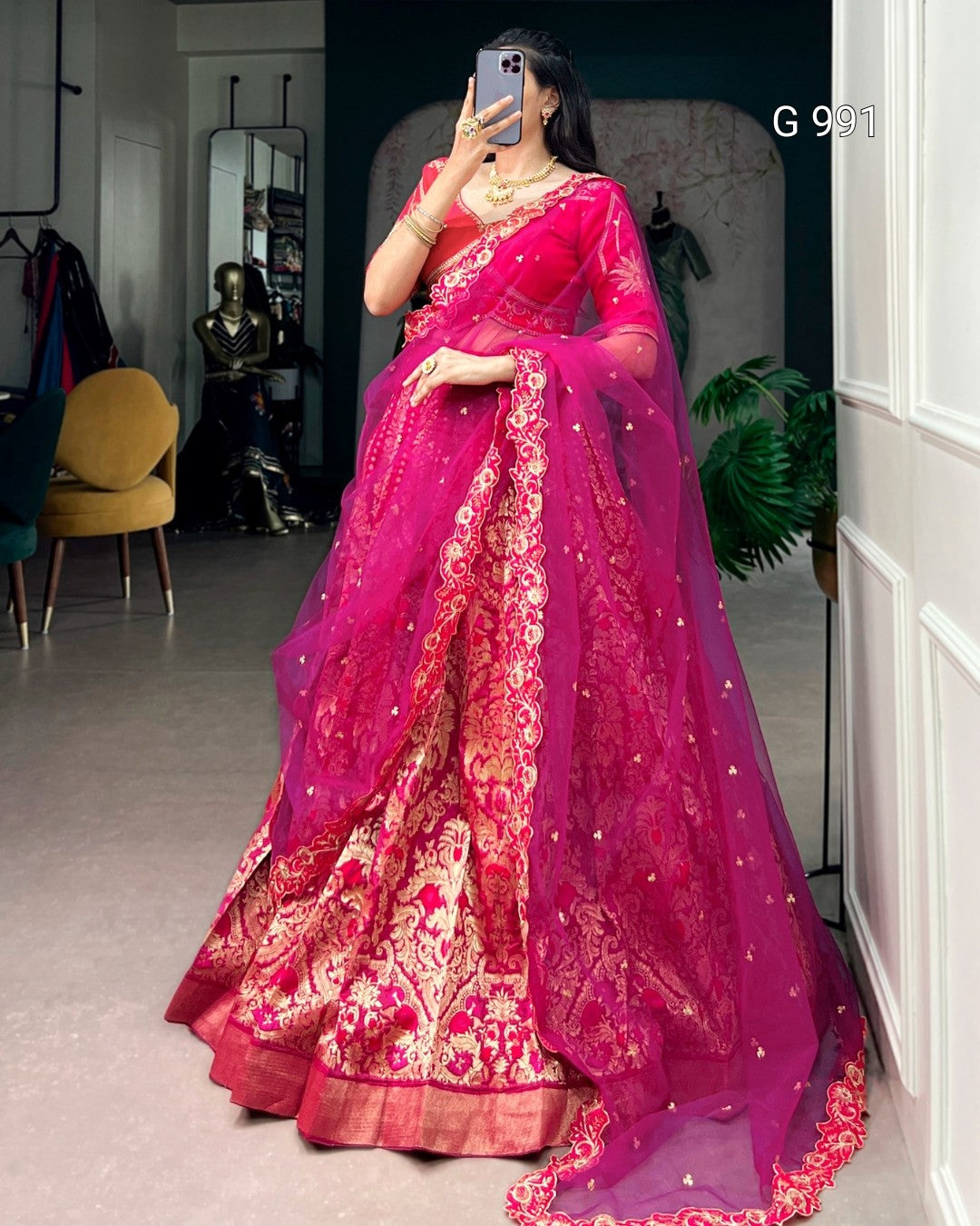 A woman takes a mirror selfie wearing a vibrant magenta pink and gold heavily embroidered traditional Indian lehenga choli set. The dress features intricate floral patterns and a matching sheer dupatta with small gold embellishments.