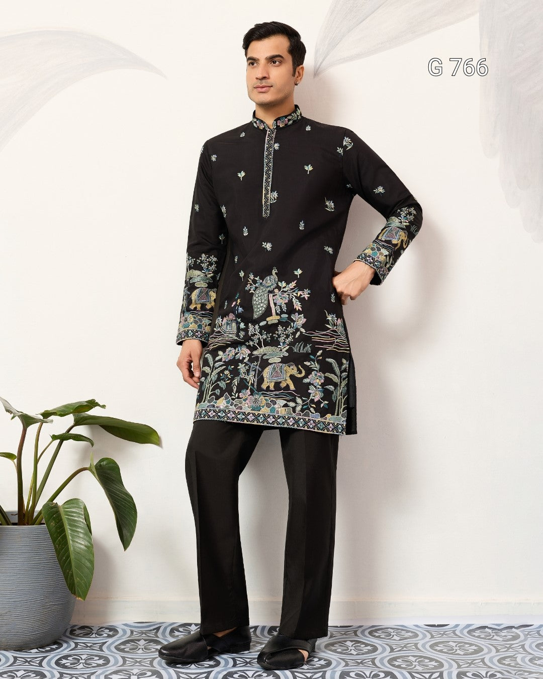 Man wearing a black embroidered kurta with floral patterns in a room with a plant and patterned floor.
