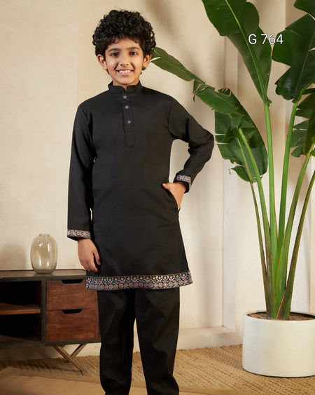 Young boy in a black traditional outfit standing indoors with a plant and wooden furniture in the background.