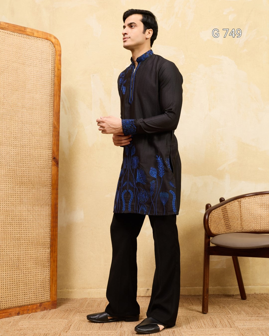 Man wearing a black kurta with blue embroidery standing against a beige wall.