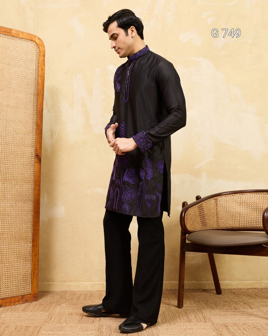 Man wearing a black kurta with purple embroidery standing in a room with a chair and wall.