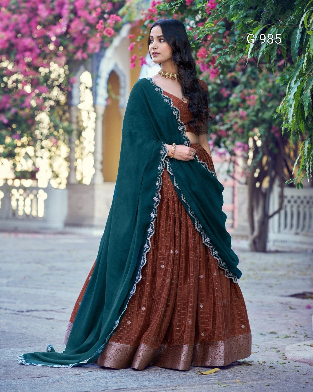 A woman wearing a traditional Indian outfit with a dark green dupatta and a brown flared skirt with metallic embroidery, standing outdoors in front of a garden with pink flowers.