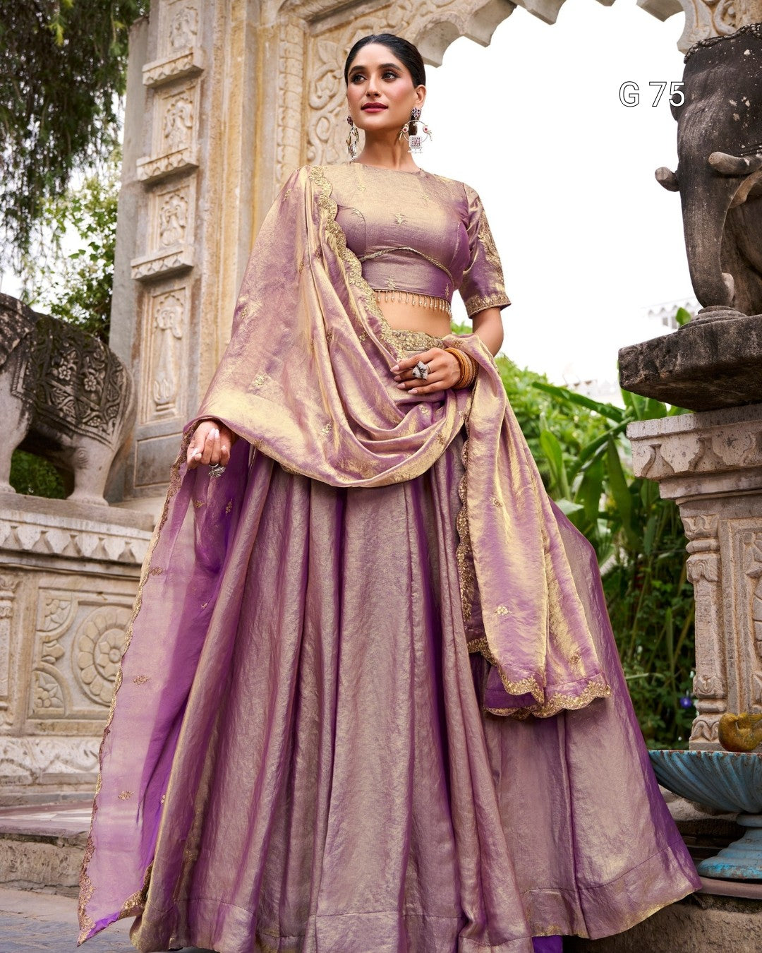 A woman models a traditional Indian outfit consisting of a lavender purple skirt (lehenga), a matching embroidered blouse, and a sheer dupatta with gold zari detailing, set against an ornate stone architectural background.
