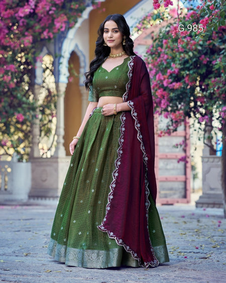 A woman models a traditional Indian ethnic wear set consisting of an olive green embroidered lehenga skirt, a matching green blouse, and a deep maroon dupatta with silver border detailing, set against a background of arches and blooming pink flowers.