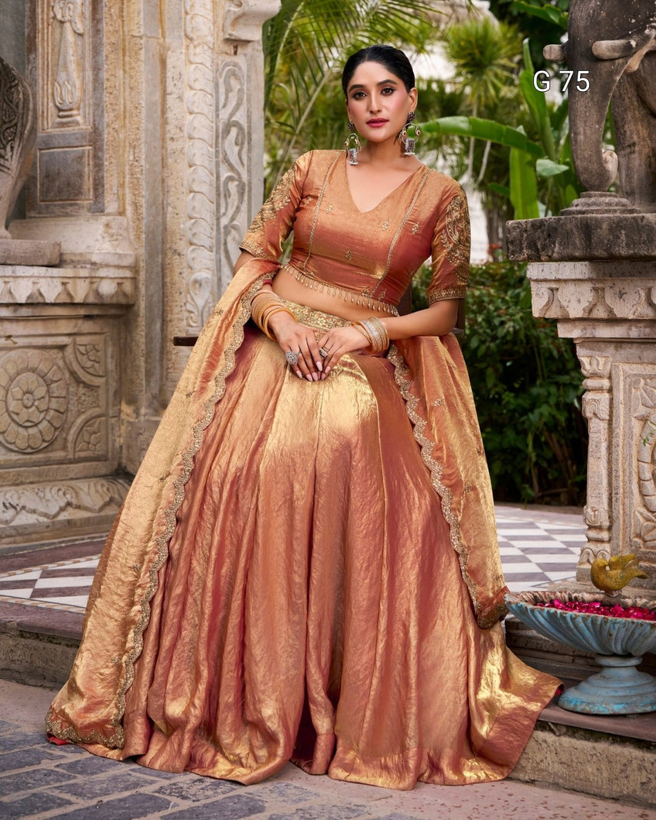 A woman models a traditional Indian salwar kameez outfit in a bronze metallic silk fabric, featuring a flared skirt, embroidered blouse, and a matching dupatta draped over her shoulder, set against an ornate stone courtyard background.