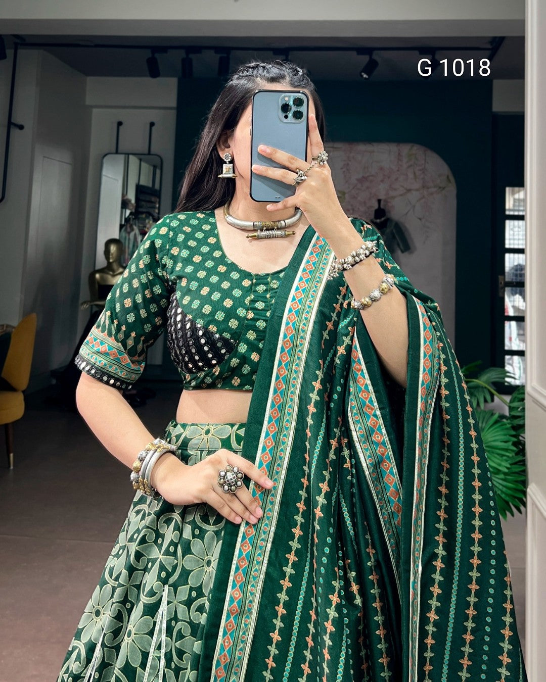 A woman takes a mirror selfie wearing a green printed traditional outfit with a matching dupatta. The outfit features a green and white patterned blouse and skirt, with a dark green dupatta decorated with a vertical border of orange, white, and green motifs.