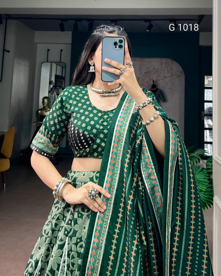 A woman takes a mirror selfie wearing a green printed traditional outfit with a matching dupatta. The outfit features a green and white patterned blouse and skirt, with a dark green dupatta decorated with a vertical border of orange, white, and green motifs.