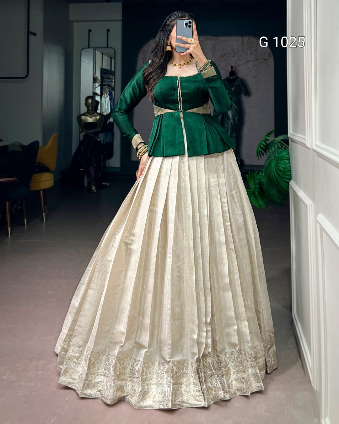 A woman takes a mirror selfie in a boutique wearing a traditional Indian outfit consisting of a dark green long-sleeve peplum top and a voluminous cream-colored flared skirt with gold zari embroidery.
