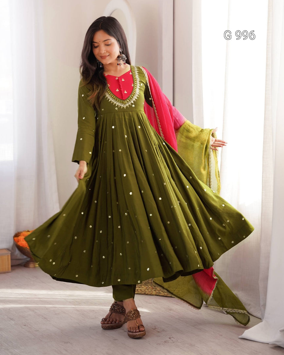 A woman modeling an olive green Anarkali gown with a red embroidered neckline, decorated with small gold dots, paired with a bright pink dupatta, in a brightly lit indoor setting.