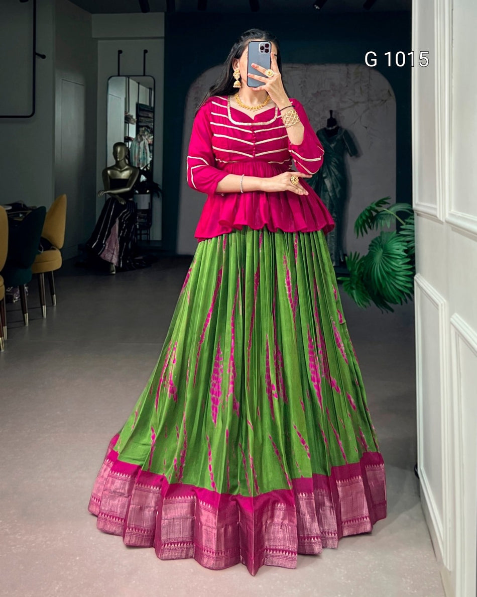 A woman takes a mirror selfie wearing a vibrant green and magenta lehenga choli set. The top is bright magenta with white stripes, and the long skirt is lime green with magenta vertical patterns and a matching magenta border.