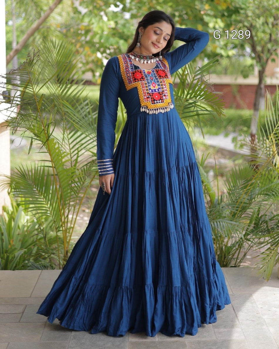 A woman wearing a long-sleeved navy blue maxi dress with colorful embroidered detailing around the neckline, standing outdoors on a tiled patio with green plants in the background.