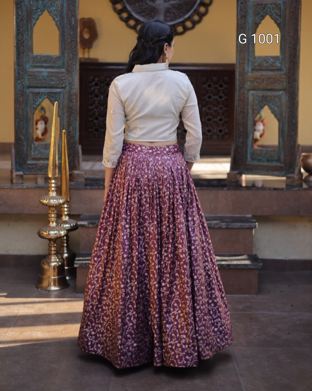 A woman viewed from behind wearing a white long-sleeve blouse and a voluminous purple floral printed skirt, standing in front of an ornate traditional doorway. The text 'G 1001' is visible in the top right corner.