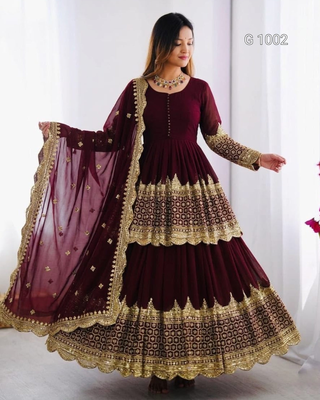 A woman modeling a deep maroon Anarkali gown with intricate gold embroidery and a matching sheer dupatta, set against a bright white indoor background.