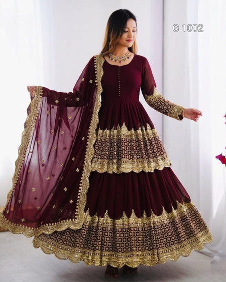 A woman modeling a deep maroon Anarkali gown with intricate gold embroidery and a matching sheer dupatta, set against a bright white indoor background.