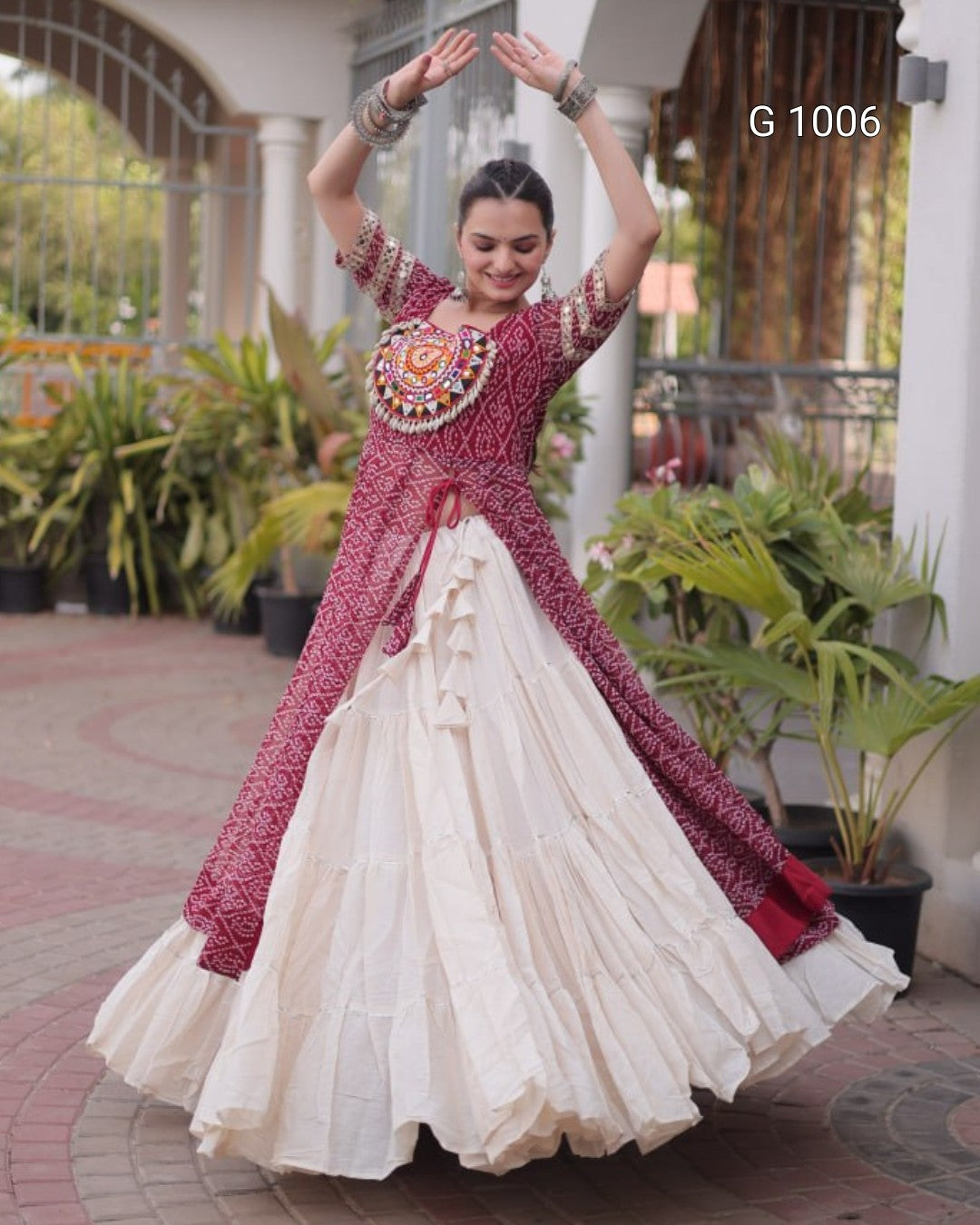 A woman modeling a traditional Indian outfit with a deep red printed kurta and a tiered cream skirt, posing outdoors in front of a white building with potted plants. The text 'G 1006' is visible in the top right corner.