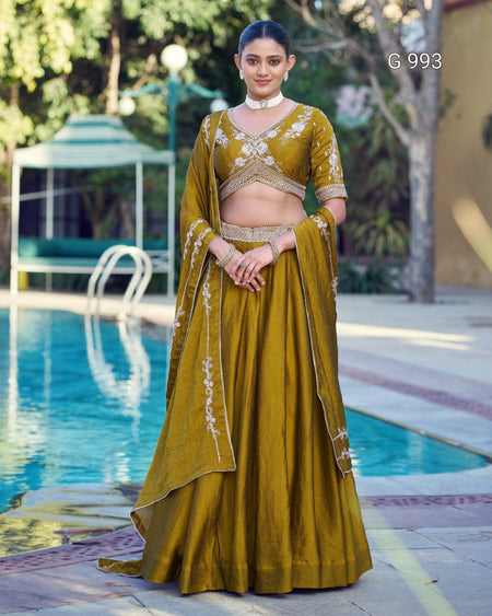 A woman stands outdoors beside a swimming pool wearing an olive yellow embroidered traditional outfit with a matching draped scarf. The outfit has ornate white and gold floral embroidery on the bodice, sleeves, and scarf.