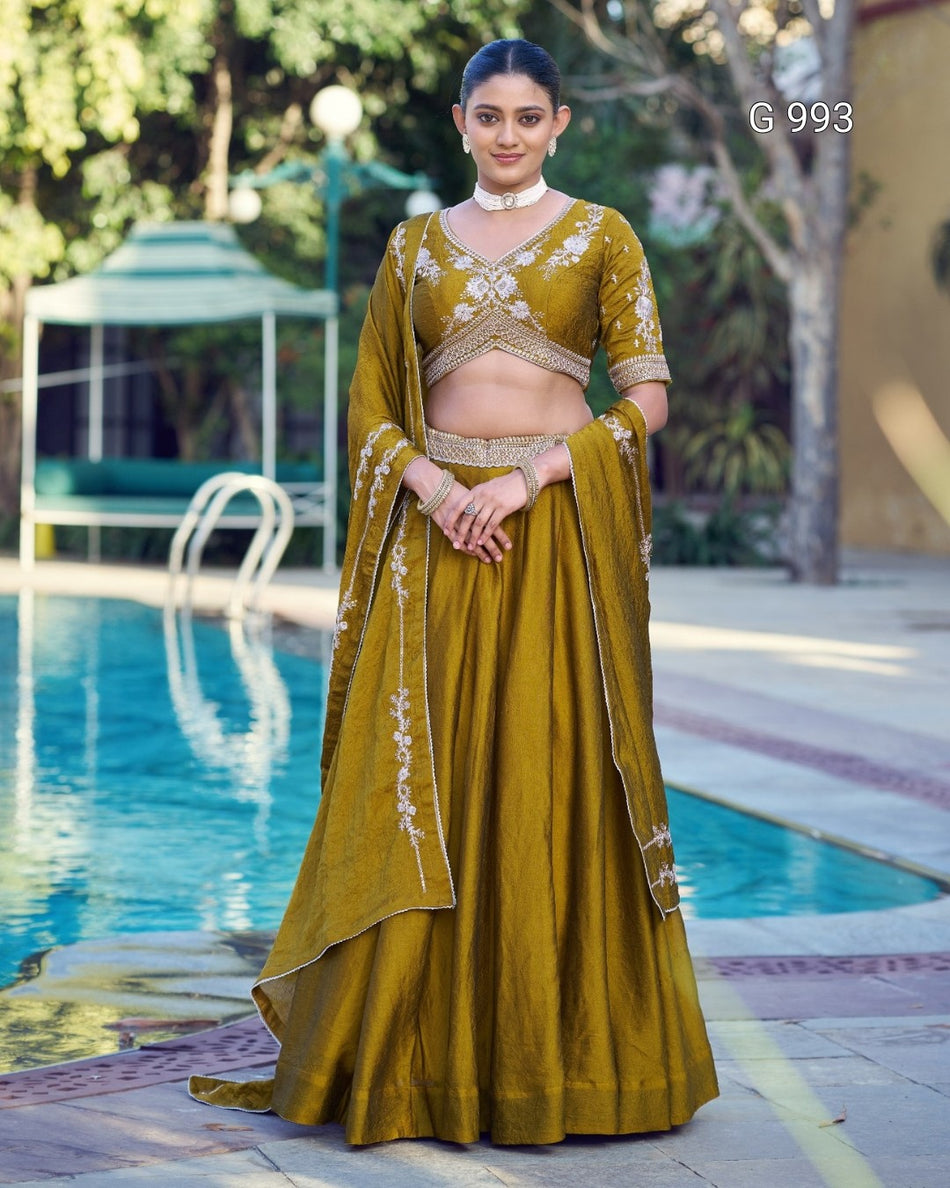 A woman stands outdoors beside a swimming pool wearing an olive yellow embroidered traditional outfit with a matching draped scarf. The outfit has ornate white and gold floral embroidery on the bodice, sleeves, and scarf.