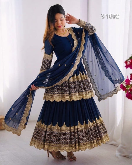 A woman modeling a navy blue traditional ethnic outfit with intricate gold embroidery and a matching sheer dupatta, posed in a bright indoor setting with white curtains.