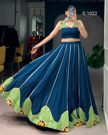 A woman modeling a two-piece traditional outfit consisting of a dark teal blue crop top and a matching flared skirt with light green border panels and gold vertical stripes. She is taking a mirror selfie in an indoor boutique setting.