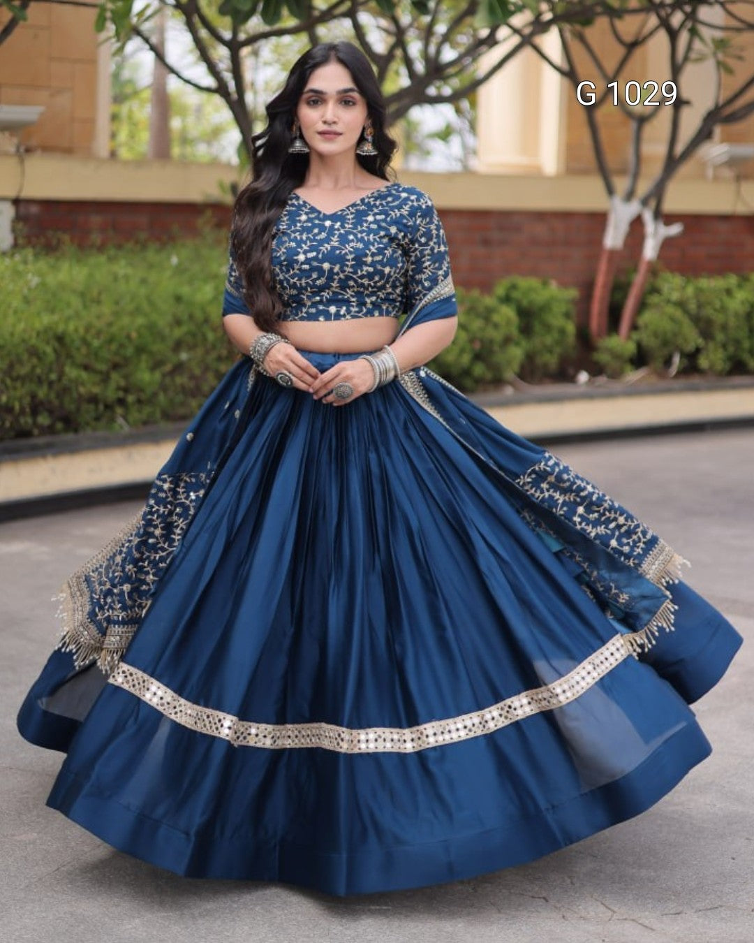 A woman modeling a navy blue traditional Indian lehenga choli set with intricate gold embroidery, standing outdoors in front of a building and trees. The set includes a heavily embroidered crop top and a voluminous flared skirt with a decorative border.