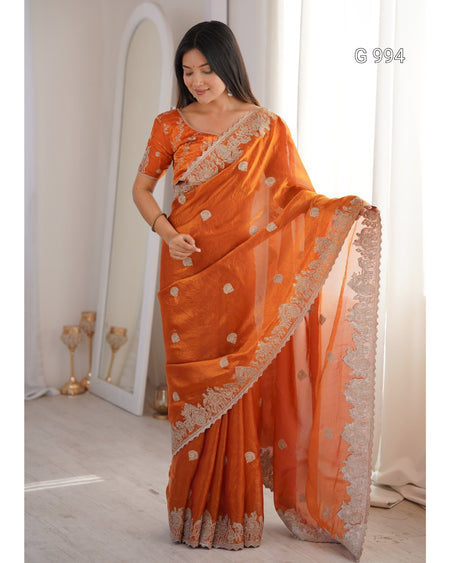 A woman models an orange saree with a matching embroidered blouse in a bright indoor setting. The saree has a sheer dupatta with a wide beige border and scattered gold floral motifs.