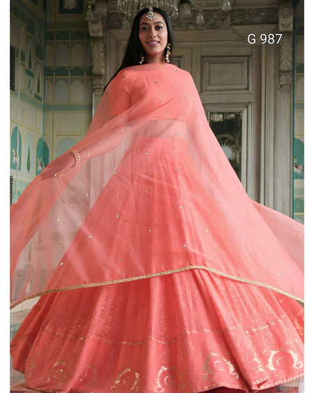 A woman models a traditional peach-pink lehenga choli set with a matching dupatta, featuring gold embroidery and mirror work, set against an ornate classical interior background.