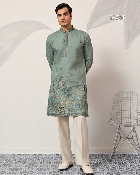 Man wearing a green embroidered kurta with white pants in a minimalistic room.