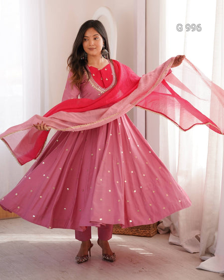 A woman modeling a pink traditional outfit with a matching dupatta, featuring a flared skirt with small silver star embellishments, in a softly lit indoor setting.