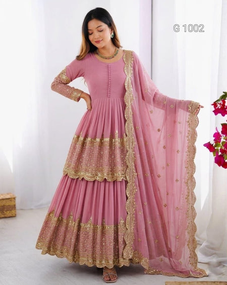 A woman models a pink traditional Anarkali gown with gold embroidery and a matching sheer dupatta, set against a white curtain background.