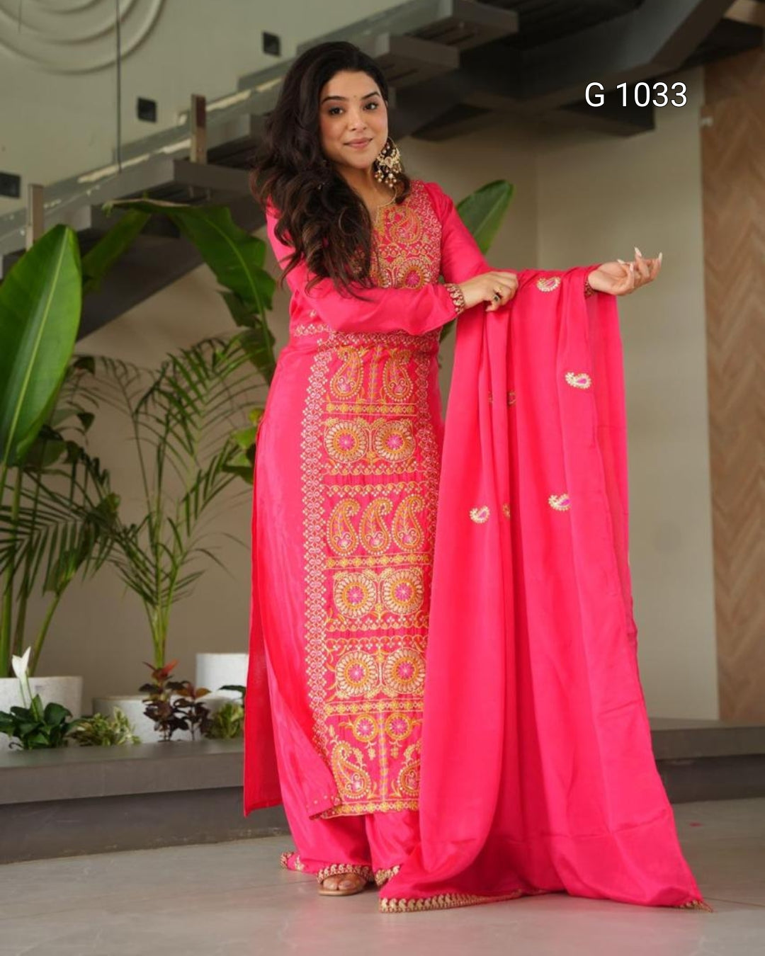 A woman modeling a bright pink traditional Indian salwar kameez suit with ornate gold embroidery, holding up the matching pink dupatta in an indoor setting with plants in the background.