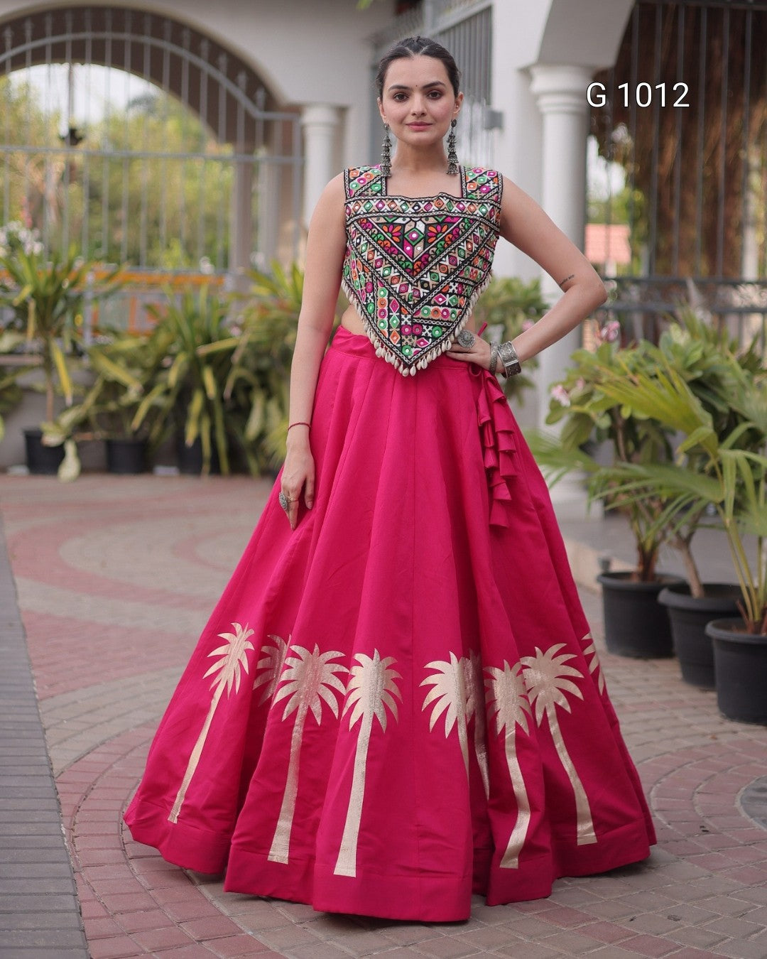 A woman modeling a bright pink traditional gown with a heavily embroidered multicolor bodice and gold palm tree motifs near the hem, standing outdoors on a paved walkway with potted plants and a building in the background.