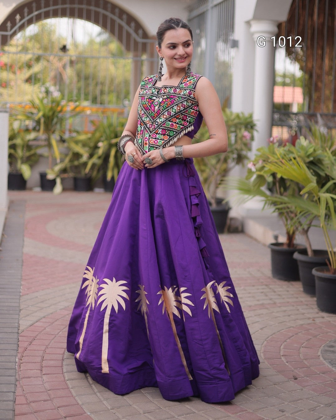A woman wearing a purple traditional formal gown with a heavily embroidered multicolor bodice and gold palm tree motifs near the hem, standing outdoors on a paved walkway with potted plants in the background.