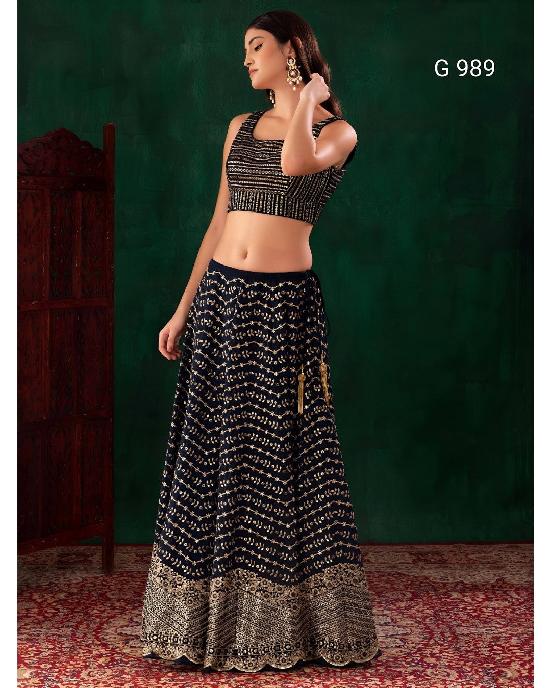 A woman modeling a black and gold patterned traditional Indian outfit consisting of a sleeveless crop top and a long flared skirt with an ornate gold border, posed against a dark green textured backdrop with a red patterned rug.