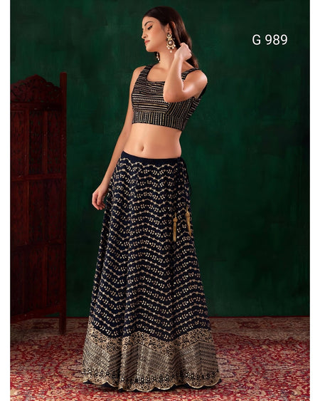 A woman modeling a black and gold patterned traditional Indian outfit consisting of a sleeveless crop top and a long flared skirt with an ornate gold border, posed against a dark green textured backdrop with a red patterned rug.