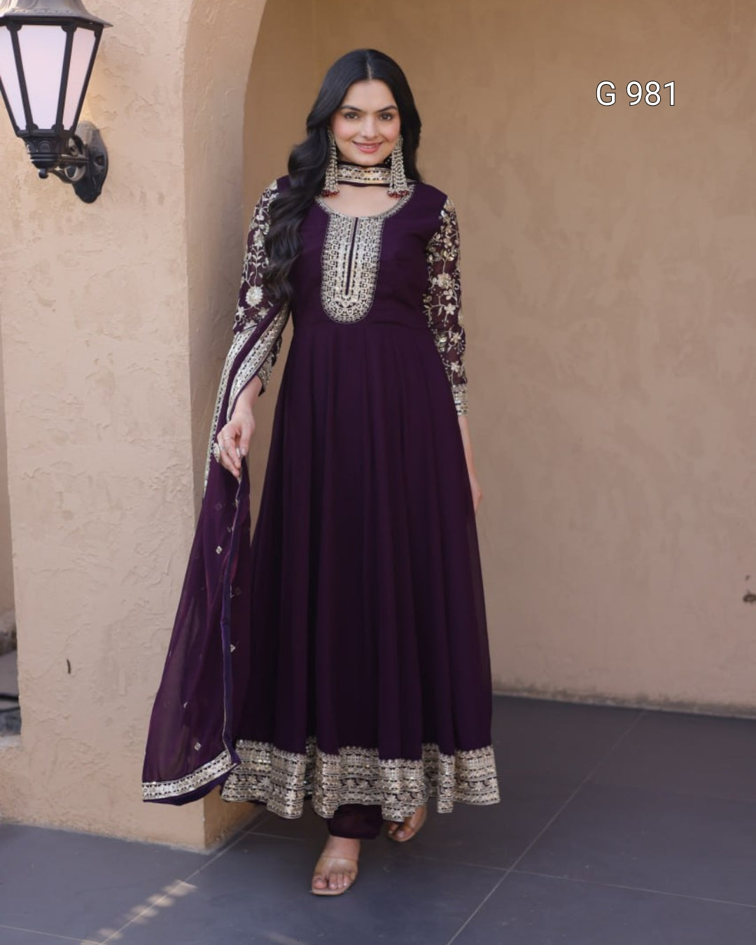 A woman models a deep purple Anarkali gown with intricate silver embroidery on the neckline, sleeves, and hem, paired with a matching sheer dupatta. She is standing in front of a beige stucco wall with a black wall lamp on the left.