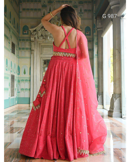A woman wearing a bright pink traditional Indian gown with gold embroidery and a matching sheer dupatta, photographed from behind in an ornate indoor corridor. The image includes the text 'G 987' in the top right.