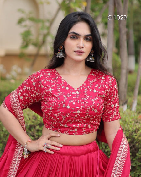 A woman modeling a bright pink traditional Indian outfit with a floral embroidered blouse and matching skirt, standing outdoors with trees in the background. The text 'G 1029' is visible in the top right corner.
