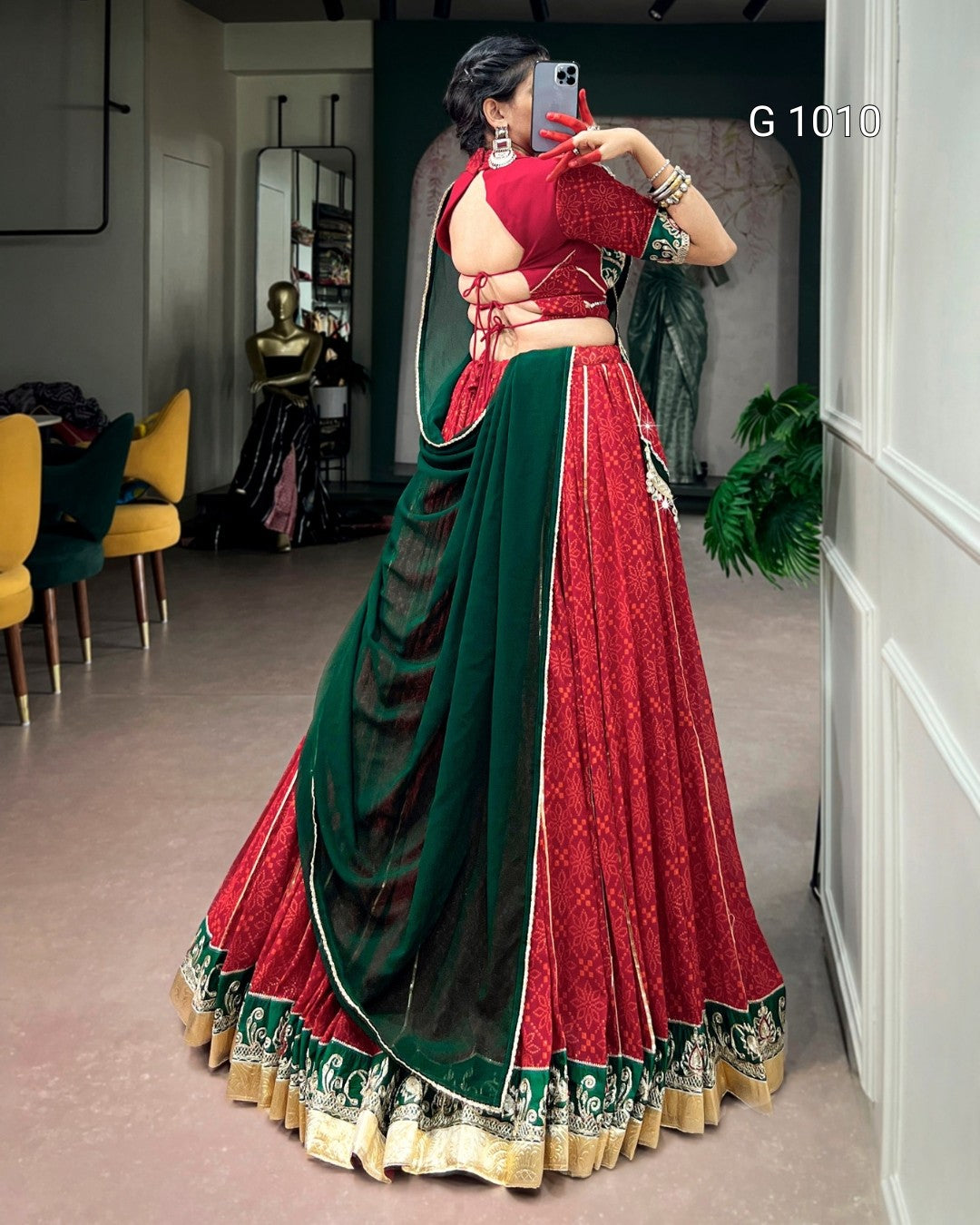 A woman takes a mirror selfie in a boutique wearing a traditional Indian lehenga choli dress. The dress features a deep red skirt and blouse with intricate gold embroidery, paired with a dark green dupatta and a matching green embroidered border on the skirt.