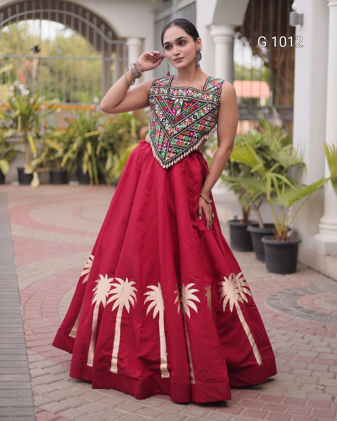 A woman models a traditional Indian salwar kameez dress with a deep red skirt featuring gold palm tree prints and a heavily embroidered multicolor kameez bodice. She is standing on a paved walkway with potted plants and a building in the background.