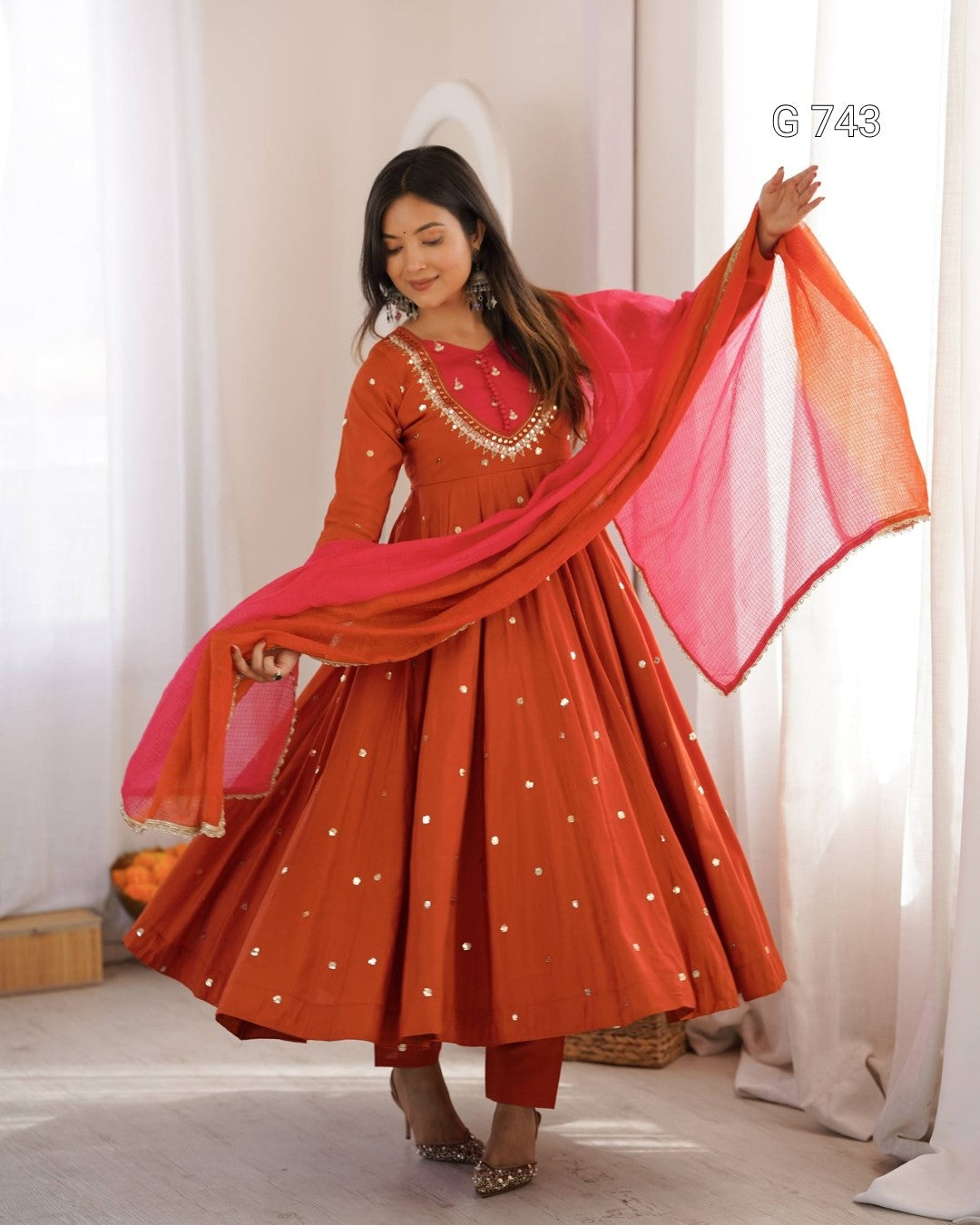 Woman in a traditional outfit with a red and pink dupatta against a white curtain background