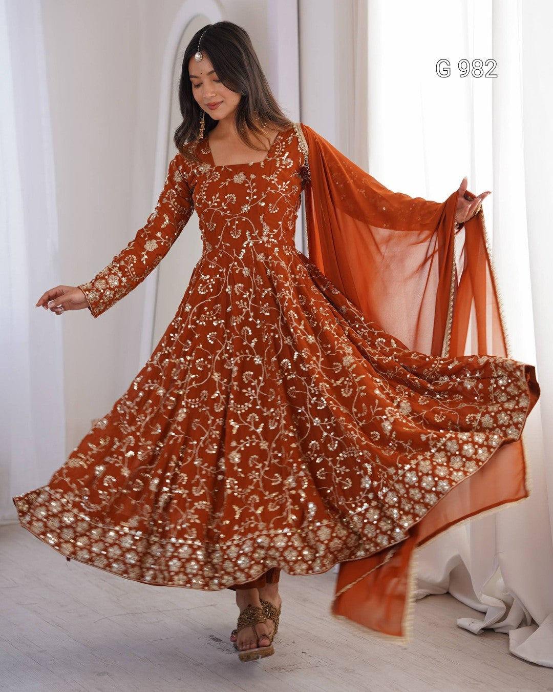 A woman models a rust-orange embroidered Anarkali gown with a matching sheer dupatta in a bright, white-walled room. The dress features intricate gold and silver floral embroidery, and the dupatta has gold border detailing.