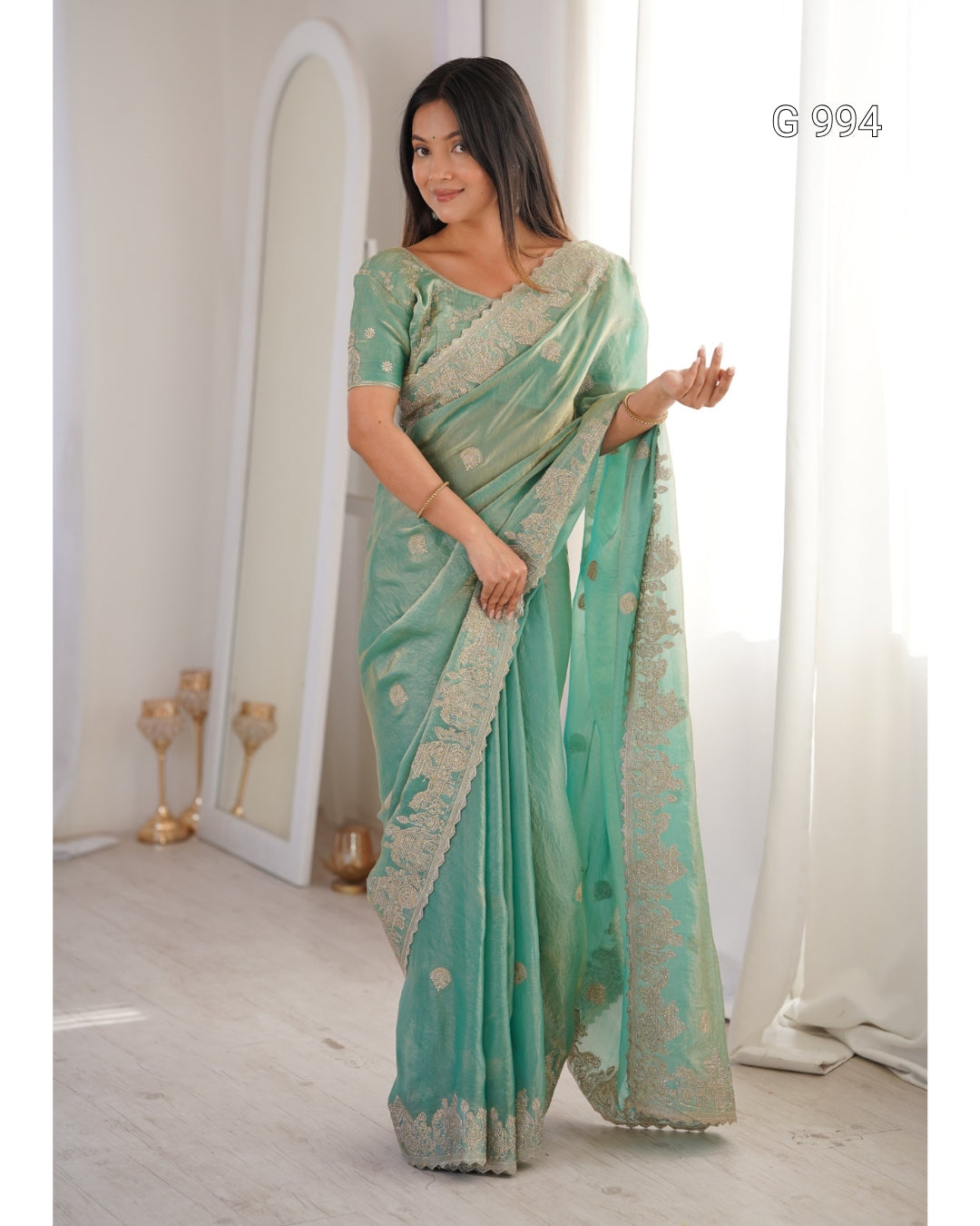 A woman modeling a mint green saree with gold floral embroidery and a wide border, standing in a bright indoor room with a mirror and white curtains. The text 'G 994' appears in the top right corner.