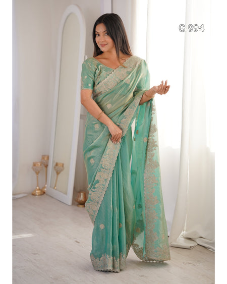 A woman modeling a mint green saree with gold floral embroidery and a wide border, standing in a bright indoor room with a mirror and white curtains. The text 'G 994' appears in the top right corner.