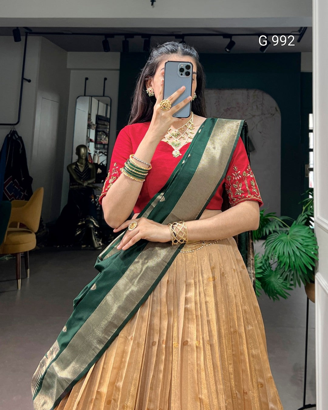 A woman takes a mirror selfie wearing a traditional Indian outfit consisting of a red embroidered blouse, a pleated beige skirt, and a dark green and gold sash. She is wearing gold jewelry and standing in an indoor room with a plant and mirror in the background.